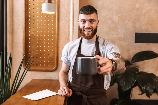 Fröhlicher Mann Barista mit Schürze steht im Café und zeigt Kaffeetasse