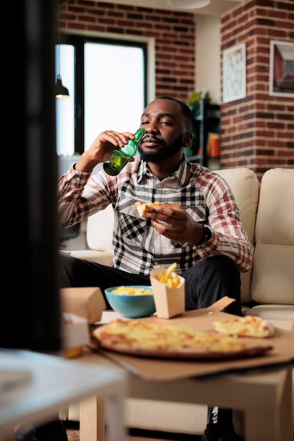 Fröhlicher Mann, der alkoholisches Bier aus der Flasche trinkt und Essen zum Mitnehmen aus dem Fast-Food-Lieferpaket isst, Film im Fernsehen sieht. Genießen Sie Getränke und Speisen zum Mitnehmen vor dem Fernseher.