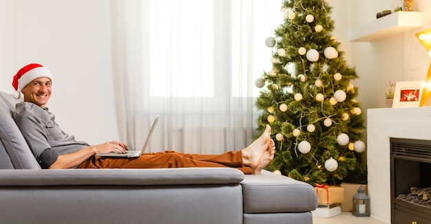 Fröhlicher Mann, der auf dem Sofa sitzt und zu Hause einen Laptop in der Nähe des Weihnachtsbaums benutzt