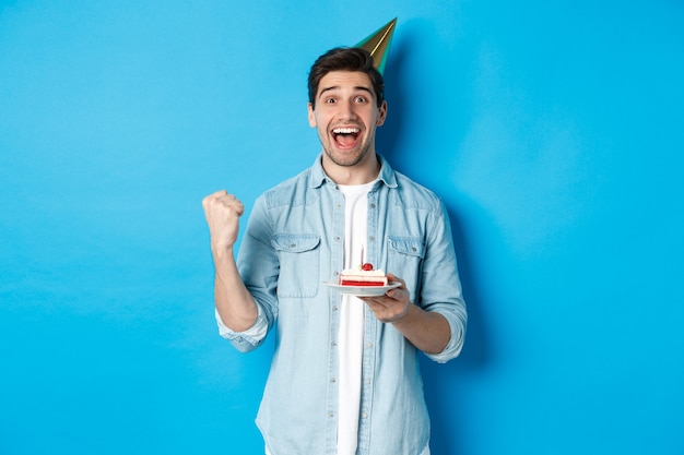 Fröhlicher Mann, der glücklich aussieht, Geburtstag im Partyhut feiert, B-Day-Kuchen hält und Faustpumpe macht, auf blauem Hintergrund stehend.