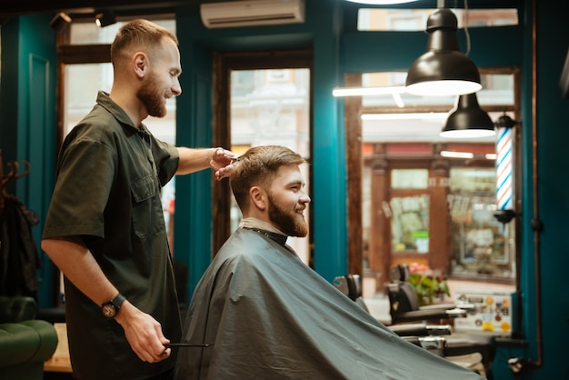 Fröhlicher Mann, der vom Friseur mit einer Schere einen Haarschnitt erhält, während er im Stuhl sitzt.