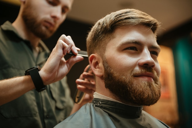 Fröhlicher Mann, der vom Friseur mit Rasiermesser einen Haarschnitt erhält, während er im Stuhl sitzt. Sieh zur Seite.