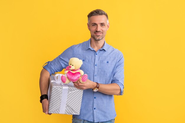 Fröhlicher Mann hält Geschenkbox und Spielzeug auf gelbem Hintergrund Valentinstag