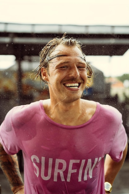 Fröhlicher Mann in einem hellen T-Shirt mit Skateboard in einem Skatepark lacht, Wasser spritzt aus seinem Haar.