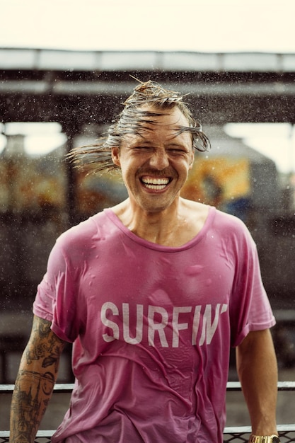 Fröhlicher Mann in einem hellen T-Shirt mit Skateboard in einem Skatepark lacht, Wasser spritzt aus seinem Haar.