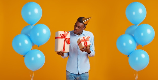 Fröhlicher Mann in Mütze mit zwei Geschenkboxen mit roten Bändern, gelber Hintergrund. Lächelnde männliche Person hat eine Überraschung, ein Ereignis oder eine Geburtstagsfeier, Ballondekoration