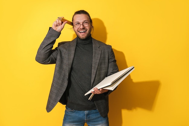 Fröhlicher Mann mit Brille, der großen Bleistift und Notizbuch auf gelbem Hintergrund hält