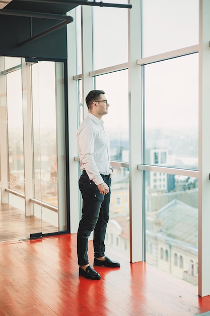 Fröhlicher Mann mit Brille und weißem Hemd steht tagsüber im Büro und schaut aus dem Fenster