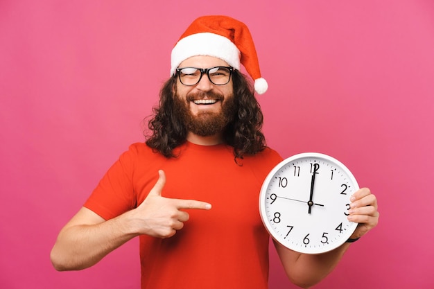 Fröhlicher Mann mit Weihnachtsmütze zeigt auf eine runde Uhr, die zwölf zeigt