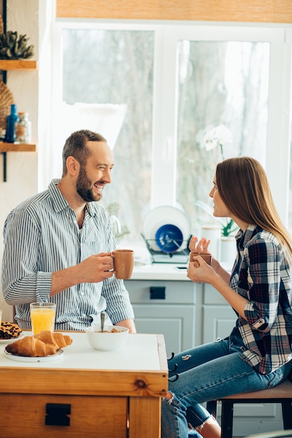 Fröhlicher Mann und Frau, die Tassen halten, während sie in der Küche sitzen und reden
