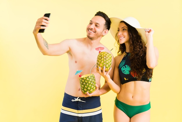 Fröhlicher Mann und Frau in Badeanzügen, die während ihrer Flitterwochen ein Selfie mit einem Smartphone machen, während sie Cocktails am Strand halten