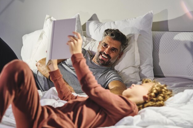 Foto fröhlicher mann und junge in schlafanzügen haben spaß, während sie auf einem weichen bett liegen und zusammen ein interessantes buch im gemütlichen schlafzimmer zu hause lesen