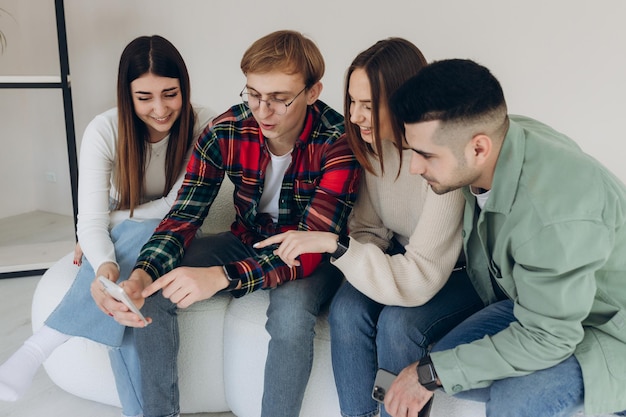 Fröhlicher Mann zeigt ein Video auf seinem Handy mit Freunden, während sie zusammensitzen und lachen Gruppe fröhlicher junger Männer und Frauen, die lachen und zu Hause auf das Smartphone schauen