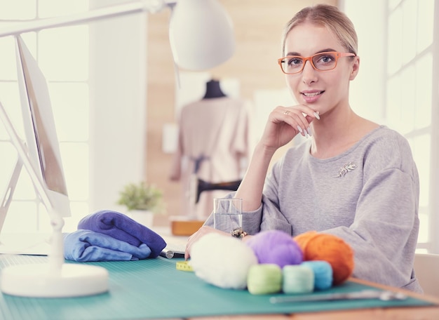 Fröhlicher Modedesigner, der in einem modernen Bürostudio mit Materialien, Stofffäden, Textillaptop auf dem Schreibtisch lächelt