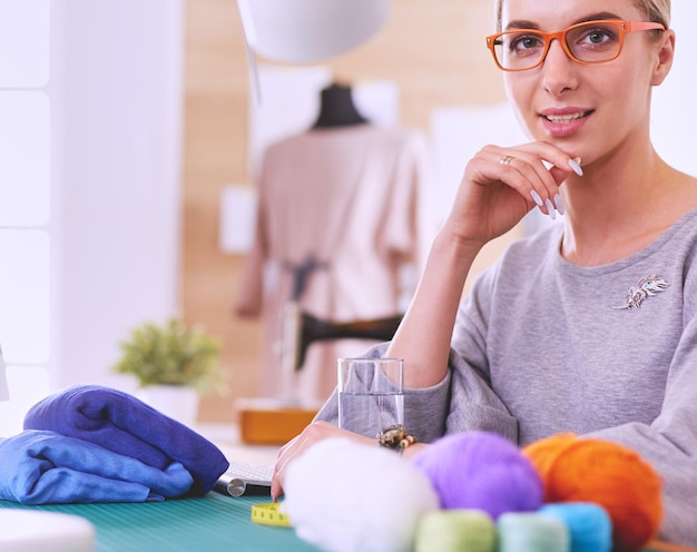 Fröhlicher Modedesigner, der in einem modernen Bürostudio mit Materialien, Stofffäden, Textillaptop auf dem Schreibtisch lächelt