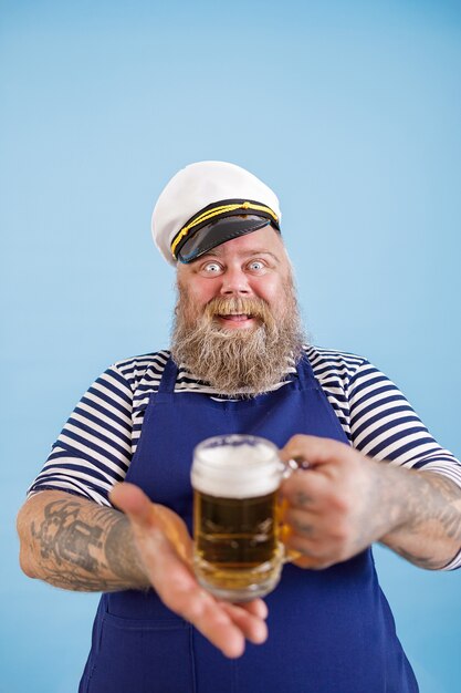 Fröhlicher molliger Mann im Matrosenkostüm bietet Glaskrug Bier auf hellblauem Hintergrund an