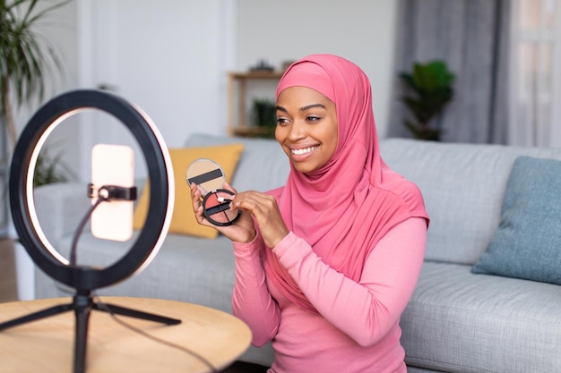 Fröhlicher muslimischer Schönheitsblogger, der Make-up-Produkte zeigt und eine Rezension macht, während er mit einem eingeschalteten Handy spricht