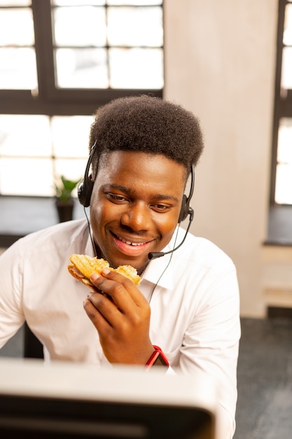 Fröhlicher netter Mann, der ein Sandwich isst, während er im Callcenter arbeitet