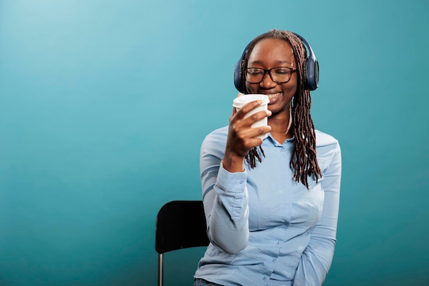 Fröhlicher positiver junger Erwachsener mit Kopfhörern, der Radio genießt, während er Kaffee trinkt. Fröhliche, glückliche afroamerikanische Frau, die herzlich lächelt, während sie auf drahtlosen Kopfhörern Musik-Playlists hört.