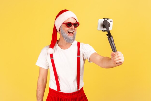 Fröhlicher positiver Mann mit stylischer Sonnenbrille und Weihnachtsmann-Hut, der Smartphone auf Selfie-Stick hält, Videos aufzeichnet oder Fotos macht, bloggt. Studioaufnahme im Innenbereich isoliert auf gelbem Hintergrund