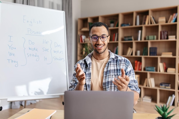 Fröhlicher professioneller männlicher Lehrer, der den Schülern per Videoanruf ein neues Thema über das Sitzen auf dem Laptop erklärt