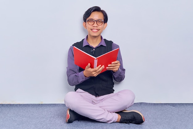 Fröhlicher Profi mit Brille, der Bericht studiert Junger asiatischer Mann trägt ein Hemd, das ein Buch hält, während er mit gekreuzten Beinen isoliert auf weißem Hintergrund auf dem Boden sitzt