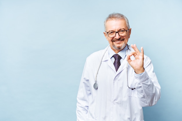 Fröhlicher reifer Arzt, der in Kamera, Gesundheitswesen und Medizin posiert und lächelt Doktor zeigt ein OK