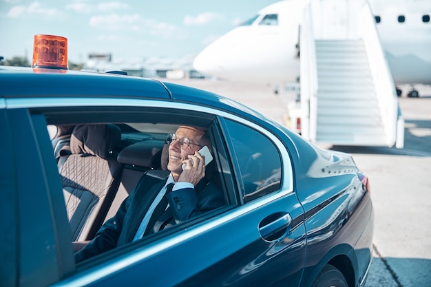 Fröhlicher reifer Geschäftsmann sitzt hinten im Auto und telefoniert beim Verlassen des Flughafens nach der Landung