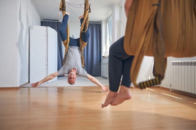 Fröhlicher reifer Yogi, der eine Inversionsübung in einer Seidenhängematte in einem Fitnessstudio durchführt