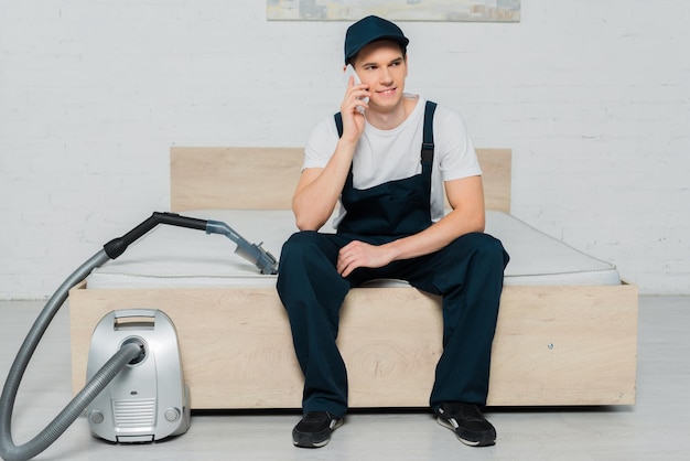 Fröhlicher Reiniger mit Mütze, der auf dem Bett sitzt und in der Nähe des Staubsaugers mit dem Smartphone spricht