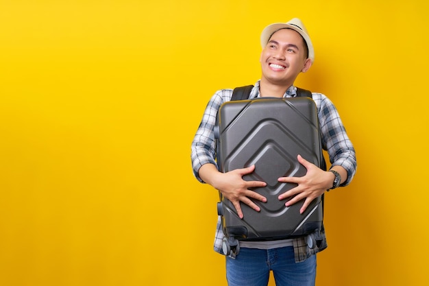 Foto fröhlicher reisender tourist asiatischer mann in freizeitkleidung hut rucksack umarmt koffertasche beiseite isoliert auf gelbem hintergrund passagierreisen ins ausland wochenenden kurzurlaub flugreisekonzept