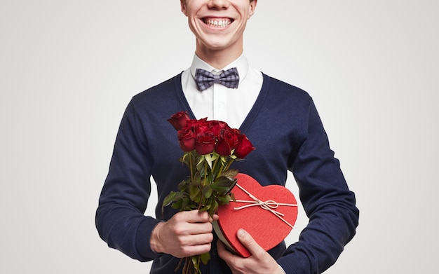 Fröhlicher romantischer Mann in elegantem Outfit mit Fliege mit Strauß roter Rosen und herzförmiger Geschenkbox zum Valentinstag