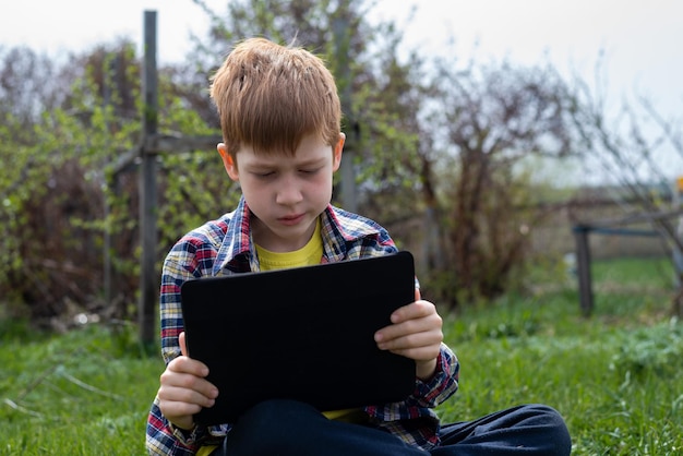 Fröhlicher rothaariger Junge, der auf dem Tablet spielt, während er auf dem grünen Gras im Hinterhof im Dorf sitzt