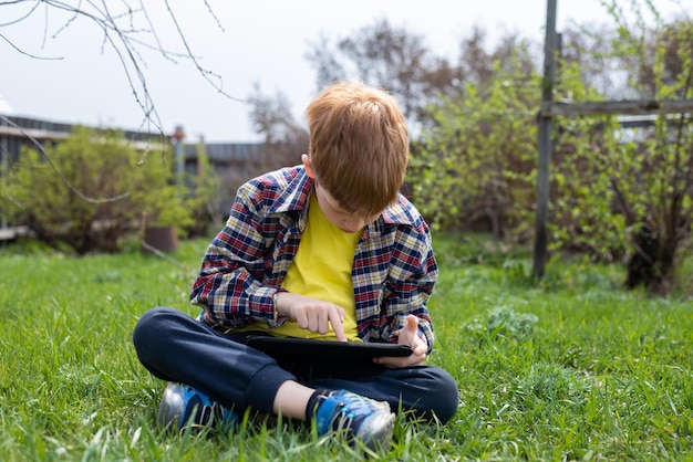 Fröhlicher rothaariger Junge, der auf einem Tablet spielt, das auf grünem Gras im Hinterhof des Dorfes sitzt
