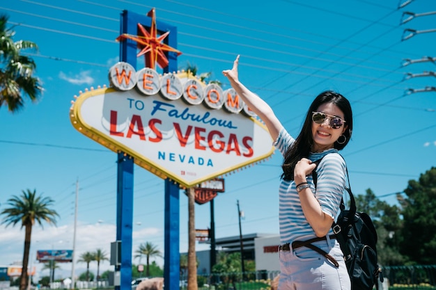 Fröhlicher Rucksacktourist des jungen Mädchens mit Sonnenbrille, der Friedensgeste zeigt und in die Kamera schaut, während er an der berühmten Werbetafel in Nevada steht. lächelnde dame reisende, die bild mit las vegas-zeichen auf sommer macht