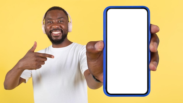Fröhlicher schwarzer Kerl mit Kopfhörern, der einen riesigen Smartphone-Gelbhintergrund zeigt