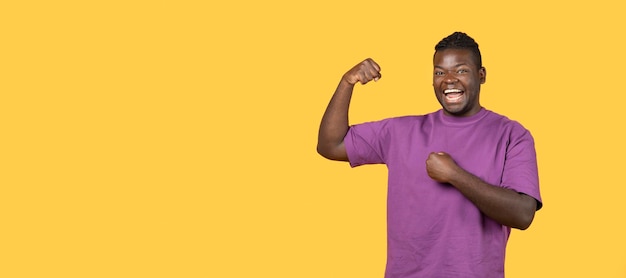 Fröhlicher schwarzer Kerl zeigt Bizepsmuskeln und gestikuliert auf gelbem Hintergrund