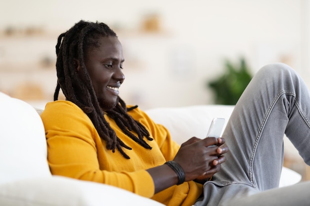 Fröhlicher schwarzer Mann, der auf dem Smartphone Nachrichten sendet, während er sich zu Hause auf der Couch entspannt