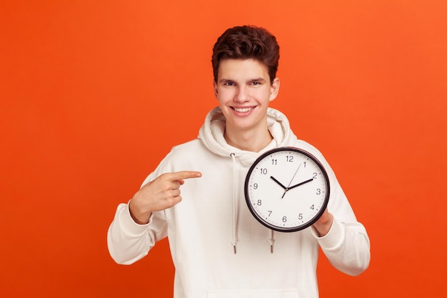 Fröhlicher, selbstbewusster junger Mann in lässigem Sweatshirt mit Kapuze, der mit dem Finger auf die Uhr in seinem Arm zeigt, Zeit, um den Start zu starten Studioaufnahme im Innenbereich isoliert auf orangefarbenem Hintergrund