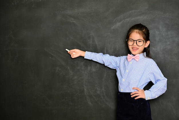 Fröhlicher, selbstbewusster Kinderlehrer, der Kreide verwendet, die auf einen leeren Bereich zeigt und das Schulkonzept zeigt, das im Hintergrund der Tafel steht.