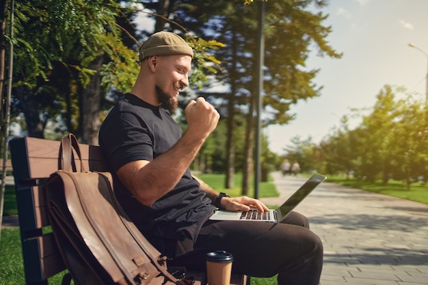 Fröhlicher siegreicher Millennial sitzt auf der Bank im Freien und schaut auf den Laptop und gestikuliert ja gewinnen Gewinner