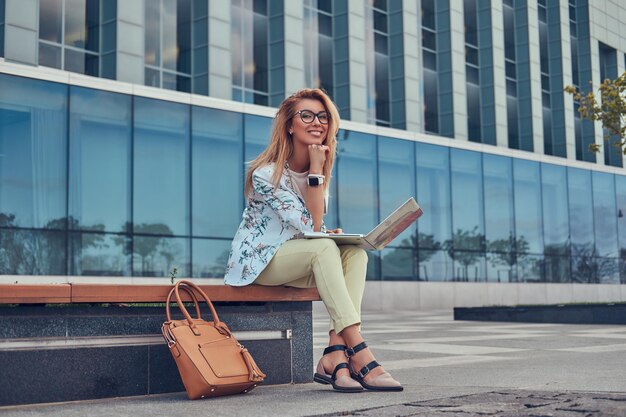 Fröhlicher, stilvoller Modeblogger, der sich im Freien entspannt, am Laptop arbeitet und auf einer Bank gegen einen Wolkenkratzer sitzt.