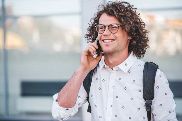 Fröhlicher, stylischer Mann mit lockigem Haar in trendiger Brille, der draußen steht, während er den Rucksack lächelnd hält und mit seinem Smartphone mit seiner Freundin auf der Stadtstraße spricht Menschentechnologiekonzept