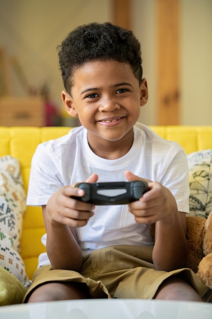 Fröhlicher süßer kleiner Junge der afrikanischen Ethnizität mit Joystick mit Lächeln, während Videospiel auf der Couch in der Freizeit palying