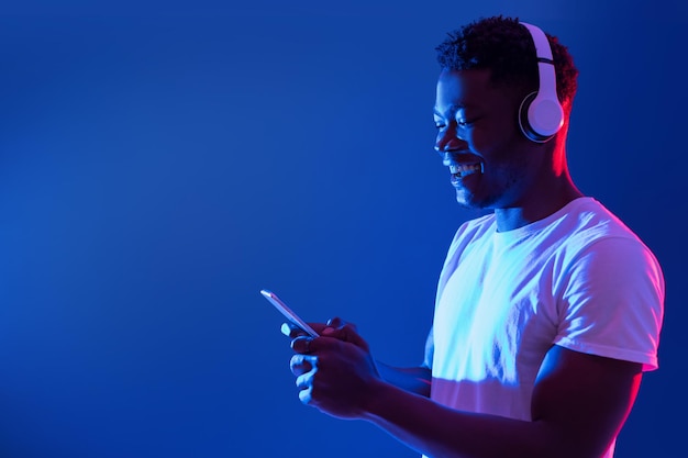 Fröhlicher tausendjähriger schwarzer Kerl in drahtlosen Kopfhörern mit Smartphone im blauen Neonlicht-Kopienraum