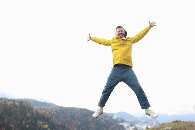 Fröhlicher Tourist im gelben Mantel lächelt und springt mit offenen Armen in die Berge und in den Wald