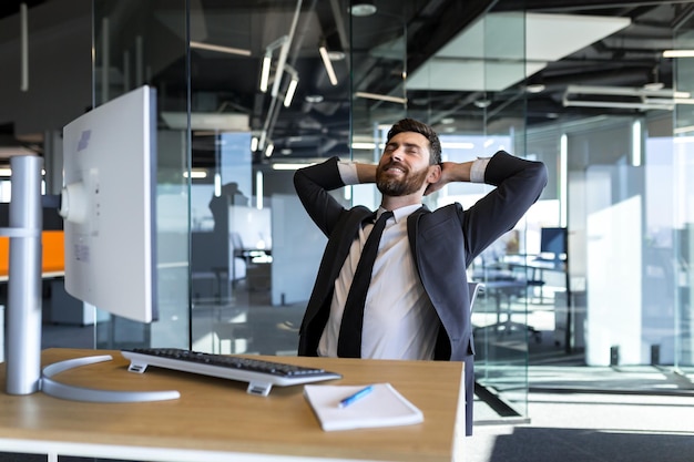 Fröhlicher und verträumter Geschäftsmann, der sich bei der Arbeit mit den Händen hinter dem Kopf ausruht und in einem modernen Büro am Computer arbeitet