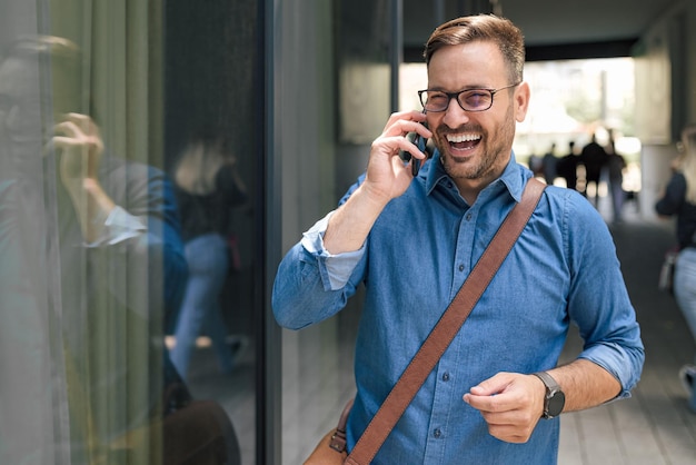 Fröhlicher Unternehmer, der auf dem Smartphone spricht und dabei über das Fenster in der Stadt nachdenkt