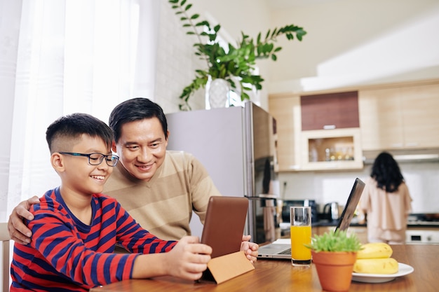 Fröhlicher vietnamesischer Vater und Sohn, die Bildungsvideo auf Tablet-Computer ansehen, wenn Mutter Abendessen kocht