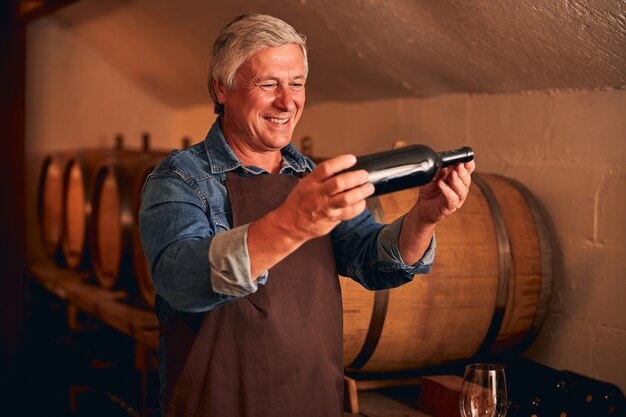 Fröhlicher Winzer in Schürze, der eine Flasche alkoholisches Getränk hält und lächelt, während er Zeit in der Weinlagerung verbringt?